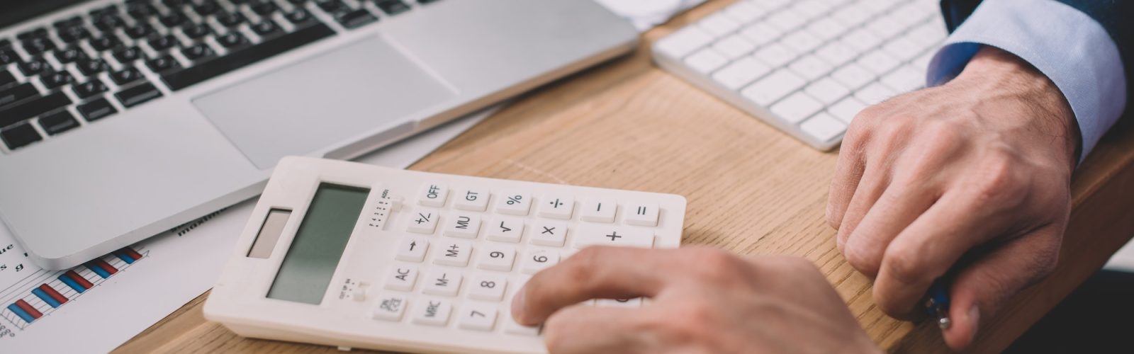 Cropped view of data analyst using calculator near laptop and papers on table